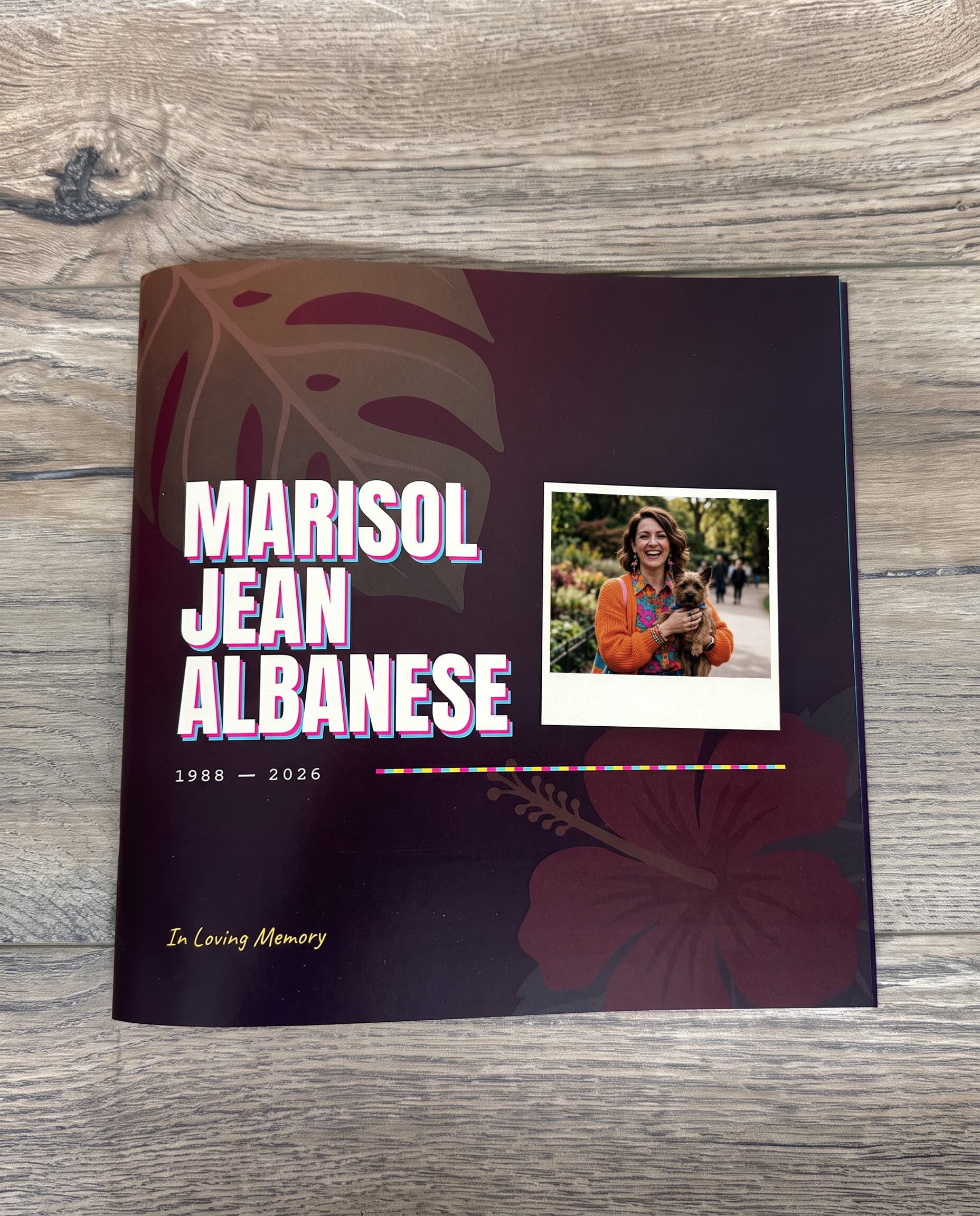 A printed memorial book on a wooden table, its dark cover patterned with tropical leaves